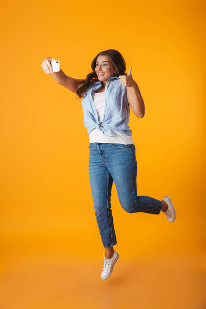 Full length of a cheerful young woman jumping isolated over yellow background, taking a selfie with outsretched hand - Fotoğraf, Görsel
