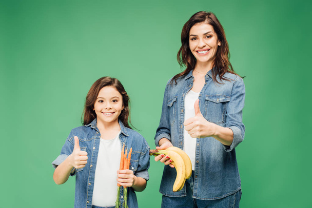 mother and daughter showing thumbs up  and holding bananas with carrots isolated on green - Fotografie, Obrázek
