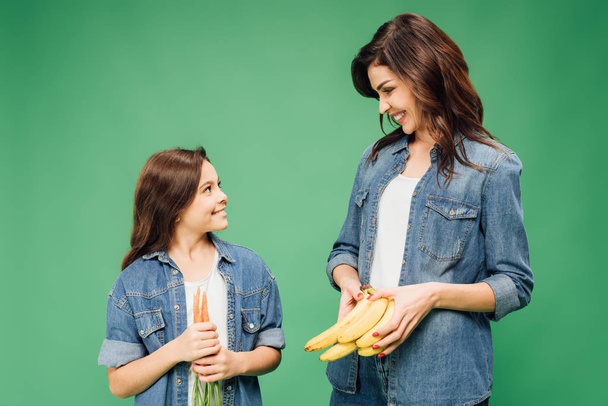 smiling mother and daughter holding bananas and carrots isolated on green - Fotografie, Obrázek