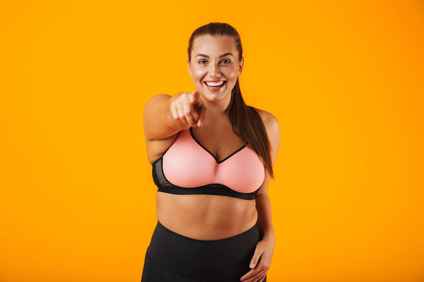 Portrait of pretty chubby woman in sportive bra smiling and standing isolated over yellow background - Fotografie, Obrázek
