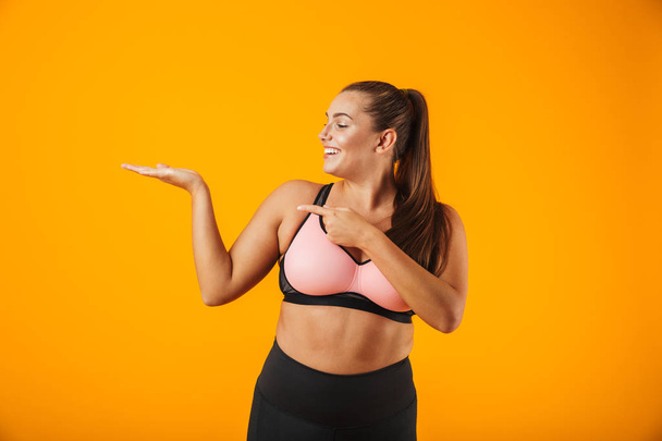 Portrait of fitness chubby woman in sportive bra holding copyspace on palm isolated over yellow background - Fotó, kép