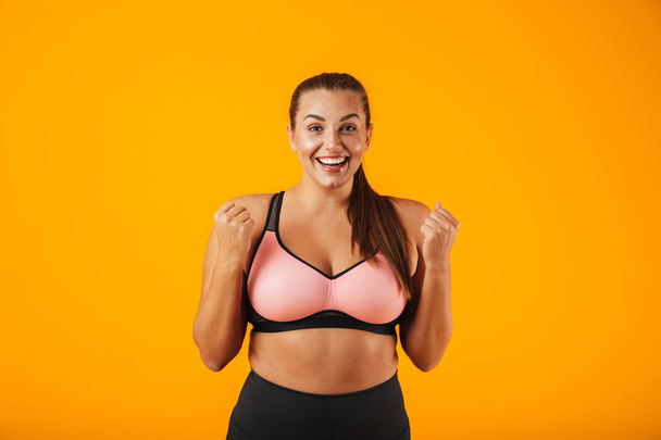 Portrait of happy chubby woman in sportive bra smiling and standing isolated over yellow background - 写真・画像