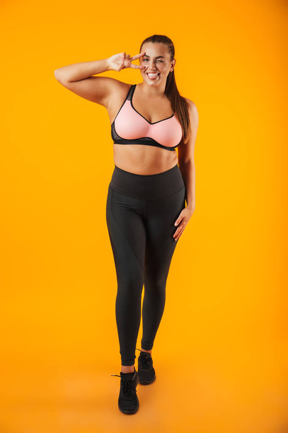 Full length portrait of beautiful chubby woman in sportive bra smiling and standing isolated over yellow background - Fotó, kép