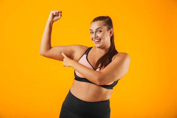 Portrait of a cheerful overweight fitness woman wearing sports clothing standing isolated over yellow background, flexing biceps - Photo, Image