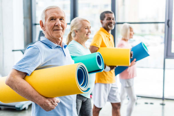 selective focus of senior sportsman holding fitness mat and his friends standing behind at gym - 写真・画像