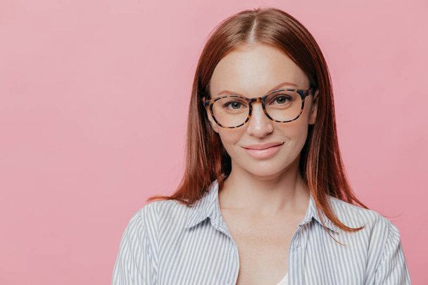 Pleased female journalist wears elegant shirt, optical glasses, has full lips, pleased to meet with people, poses against pink studio wall with blank space for your promotional content. Indoor shot - Photo, image