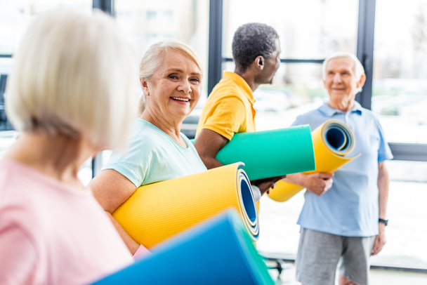 selective focus of senior sportswoman holding fitness mat and her friends standing behind at gym - Photo, Image