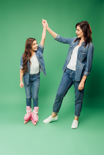 happy daughter in rollerblades cheering and holding hands with mother on green background - Fotoğraf, Görsel