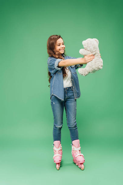 smiling child in roller blades holding teddy bear on green background - Photo, image