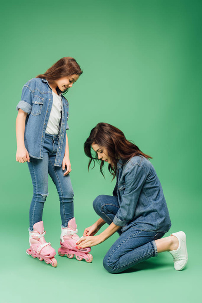mother in denim putting roller blades on daughter on green background - Фото, зображення