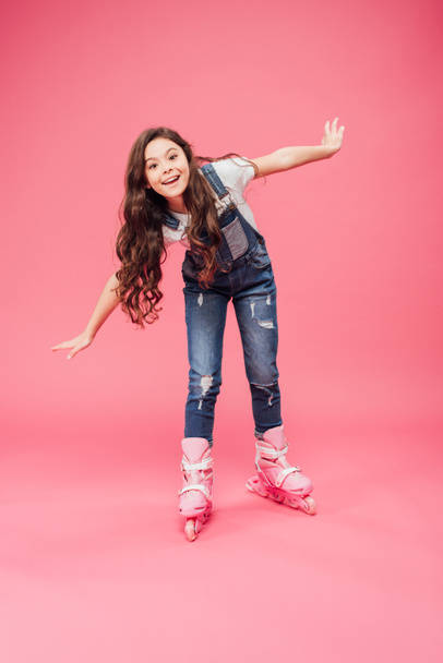 cute smiling child in overalls rollerblading with outstretched hands isolated on pink background - Fotografie, Obrázek