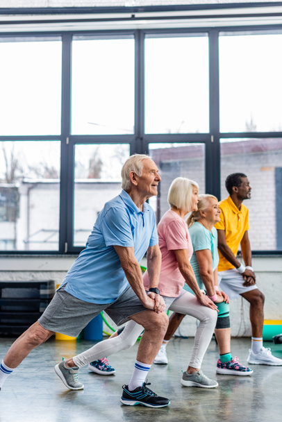 vista laterale di sportivi multietnici anziani stretching sincrono al palazzetto dello sport
 - Foto, immagini