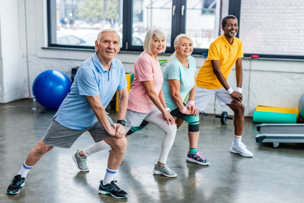 усміхнені старші багатоетнічні спортсмени синхронно розтягуються в спортивному залі
 - Фото, зображення