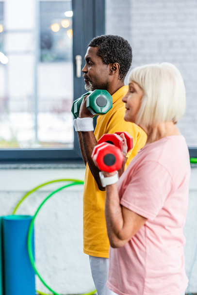 vista lateral de pareja madura interracial haciendo ejercicio con pesas en el gimnasio
 - Foto, Imagen