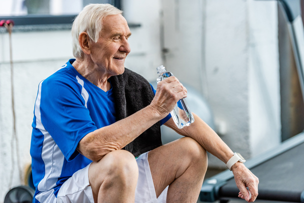 sportivo anziano sorridente con asciugamano e bottiglia d'acqua che riposa in palestra
 - Foto, immagini