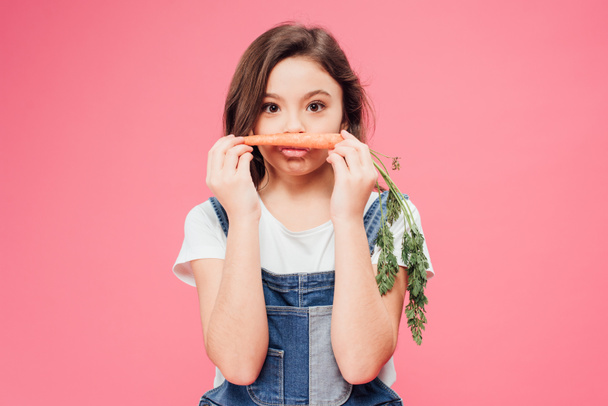 funny kid holding carrot near mouth isolated on pink  - 写真・画像