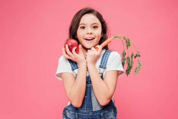 excited child holding red apple and carrot in hands isolated on pink  - Foto, Bild