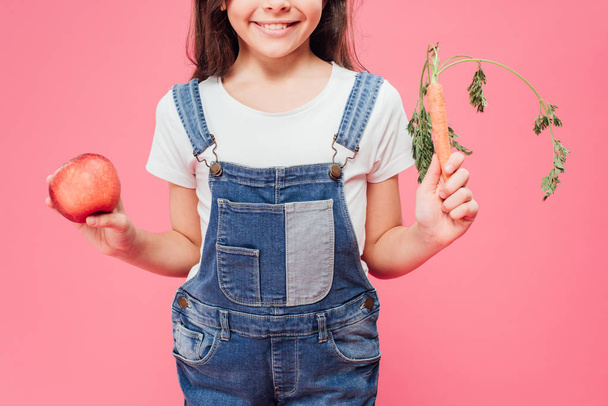 przycięte widzenia dziecko trzymając Czerwone jabłko i marchew w ręce na różowym tle  - Zdjęcie, obraz