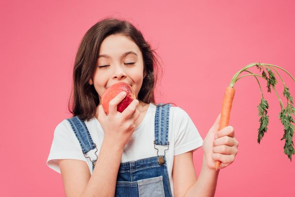 happy child eating child red apple and holding carrot isolated on pink  - Foto, immagini