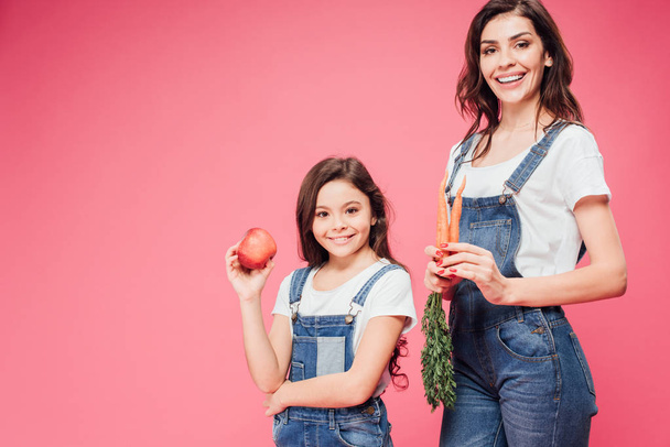 happy mom and daughter holding red apple and carrots isolated on pink  - Fotografie, Obrázek
