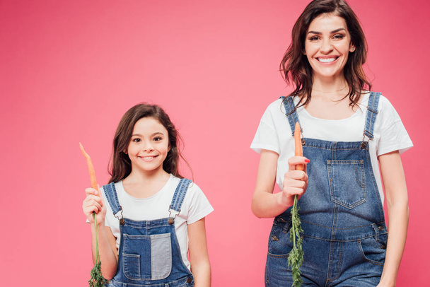 mother and daughter holding fresh carrots isolated on pink  - Foto, afbeelding