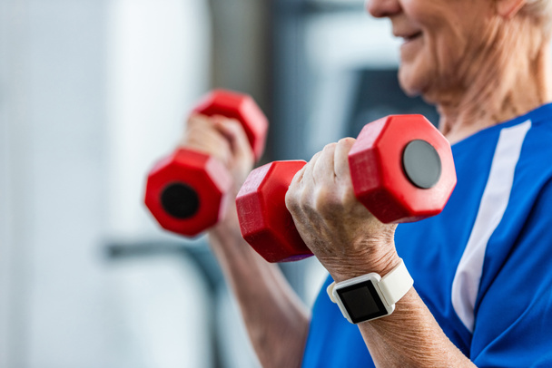 vista parcial del deportista senior con smartwatch haciendo ejercicio con pesas en el gimnasio
 - Foto, imagen