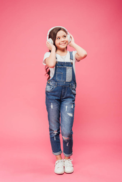 full length of cheerful kid listening music and touching headphones on pink background - Foto, Bild