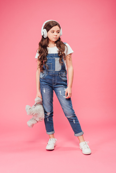 full length of sad kid standing in headphones and holding teddy bear on pink background  - Foto, afbeelding