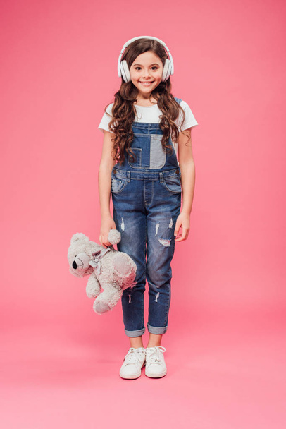 full length of smiling child standing in headphones and holding teddy bear on pink background  - Foto, Bild