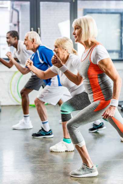 вид сбоку мультиэтнических старших спортсменов синхронно тренирующихся в спортзале
 - Фото, изображение