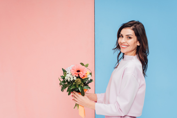beautiful woman holding bouquet and smiling at camera on blue and pink background - 写真・画像
