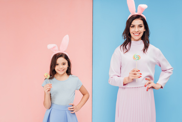 beautiful woman and child in bunny ears holding candies while looking at camera on blue and pink background - Фото, изображение