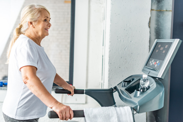side view of smiling senior sportswoman running on treadmill at sports hall - Fotó, kép
