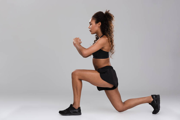 Portrait of an afro american young strong sportswoman isolated over gray background, doing lunges - Фото, изображение