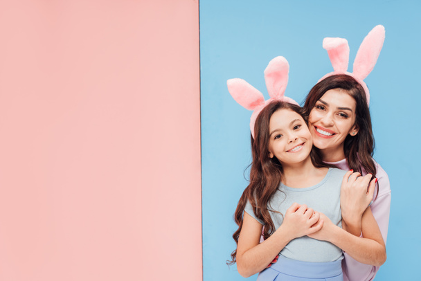 beautiful woman in bunny ears hugging child and smiling at camera on blue and pink background - Фото, изображение