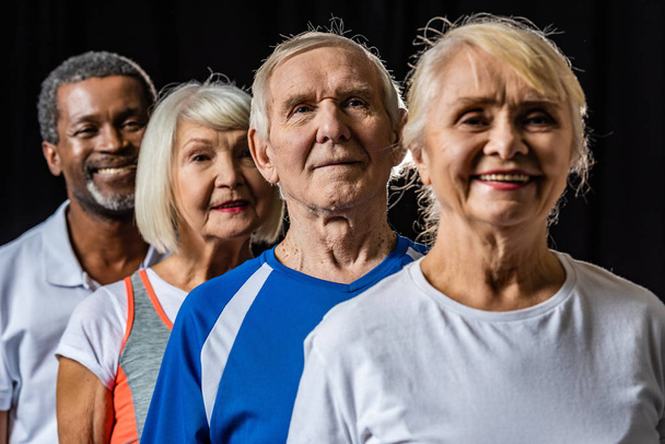 sorrindo esportistas seniores multiculturais em pé isolado em preto
 - Foto, Imagem