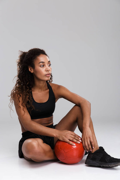 Portrait of an afro american confident young fit sportswoman doing exercises with heavy ball isolated over gray background, sitting, looking away - Foto, afbeelding