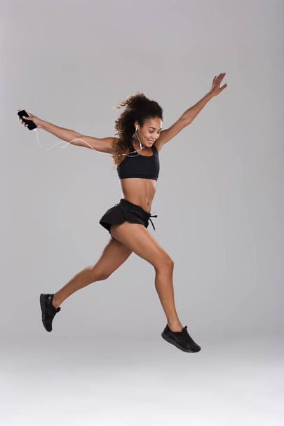 Full length portrait of an afro american young sportswoman isolated over gray background, listening to music with earphones, holding mobile phone, jumping - Photo, image
