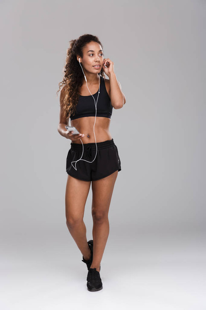 Full length of a beautiful african sportswoman isolated over gray background, listening to music with earphones, holding mobile phone - Zdjęcie, obraz