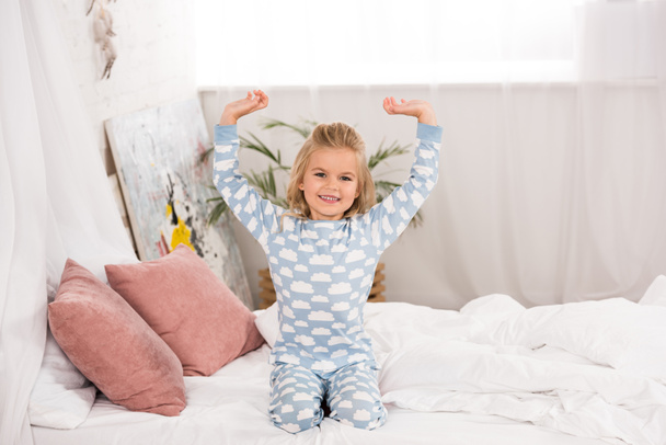 smiling cute child in pajamas sitting on bed with hands in air  - Fotografie, Obrázek