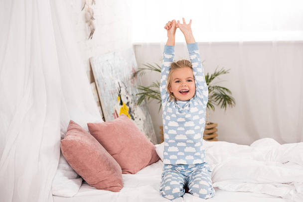 glimlachend schattig kind in pyjama zittend op bed met handen in de lucht  - Foto, afbeelding