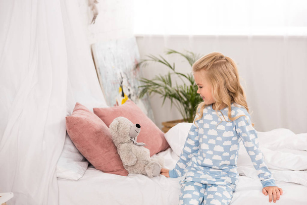 mignon adorable enfant en pyjama assis sur le lit et regardant ours en peluche
 - Photo, image