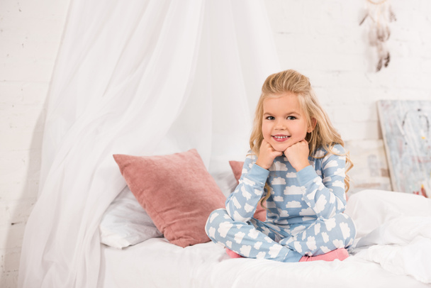 aranyos gyerek ül az ágyon a keresztezett lábak és kezek közelében arc mosolyog - Fotó, kép