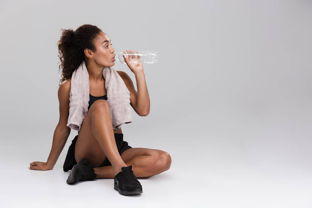 Portrait of a young african sportswoman resting after training isolated over grey background, drinking water - Fotografie, Obrázek