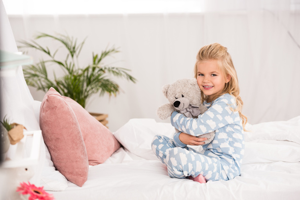 Niedliches Kind sitzt mit überkreuzten Beinen im Bett und umarmt Teddybär - Foto, Bild