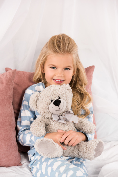 cheerful cute kid embracing teddy bear in bed - Fotó, kép