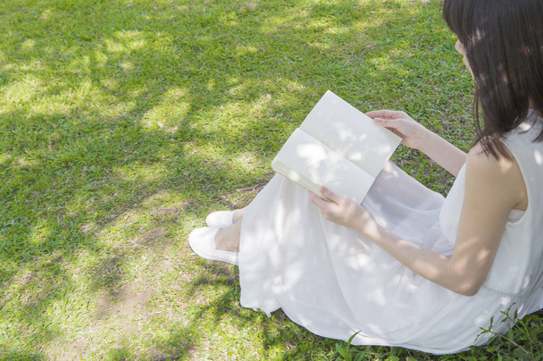 Νεαρά ασιατικές γυναίκα φοράει ένα άσπρο φόρεμα, διαβάζοντας ένα βιβλίο κάτω από το δέντρο - Φωτογραφία, εικόνα