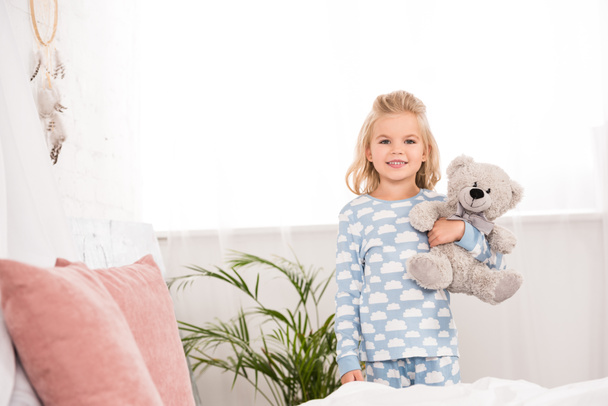 smiling child in pajamas standing with teddy bear in bedroom  - Фото, изображение