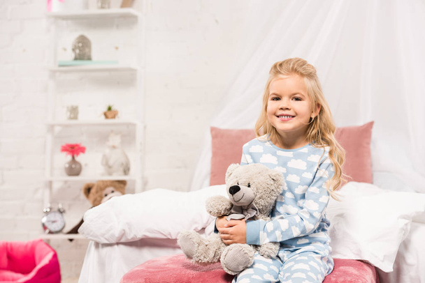 smiling cute kid sitting on bed with teddy bear and looking at camera - Photo, Image
