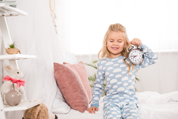 happy adorable kid sitting on bed with clock and closed eyes - Foto, Imagen